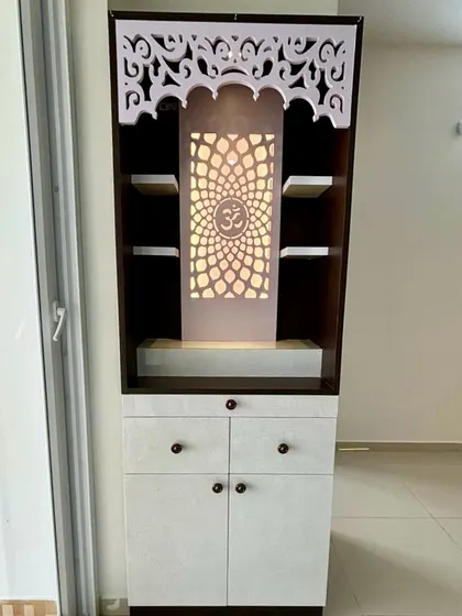 A compact and elegant pooja unit from a 2BHK project, featuring intricate white jali work, a backlit 'Om' panel, and practical storage drawers below.
