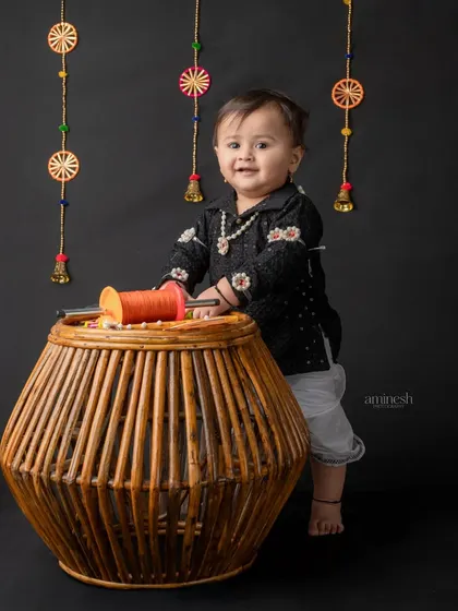 A smiling baby stands next to a basket full of kites and spools. The black background makes the child's happy expression and festive outfit the center of attention.