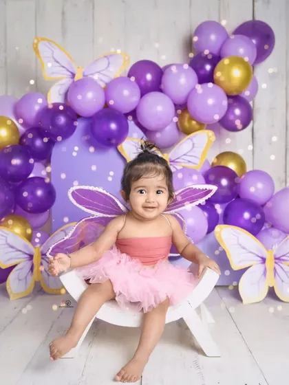 A sweet toddler dressed as a butterfly, smiling brightly for her milestone photoshoot. The purple and gold balloon arch and butterfly cutouts create a magical, whimsical setting.