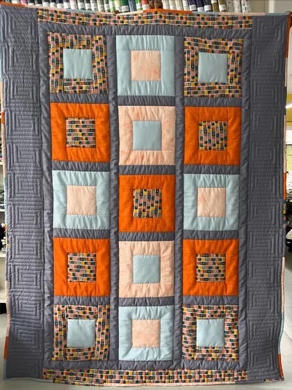 A modern take on a classic block quilt, featuring a "square in a square" design in a contemporary color palette of grey, orange, and blue.