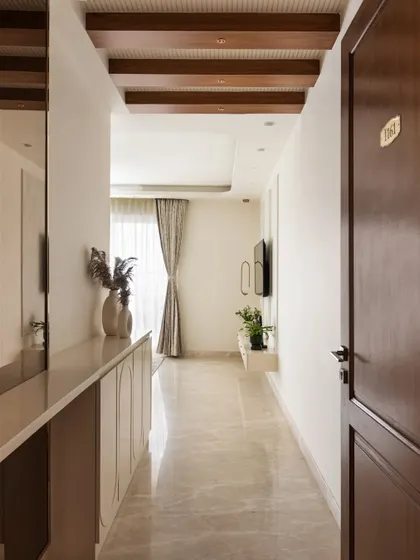 The foyer sets a tone of understated luxury. A tall mirror with a PVD golden frame adds a touch of glamour, while a clean, elegant foyer unit serves as both a console and a shoe rack. The ceiling, with its wooden rafters and grooved paneling, adds warmth and definition.