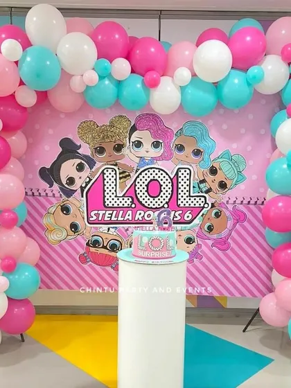 A LOL Surprise themed 6th birthday party. The decoration features a character backdrop framed by a pink, white, and teal balloon arch.