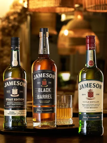 The Jameson family. This shot showcases three different Jameson expressions in a warm, inviting bar environment. The background is softly blurred to keep the focus on the distinct character of each bottle.