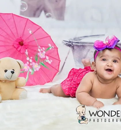 A happy baby girl lying on a fluffy white rug with a pink umbrella and a teddy bear. This simple yet adorable setup is perfect for capturing a baby's joyful spirit.