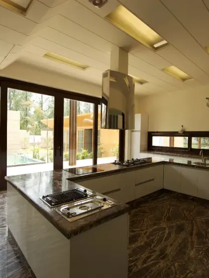 The kitchen at Sachdeva Farmhouse is a modern, light-filled space with large sliding doors opening to the poolside. Sleek white cabinetry contrasts with rich stone flooring, combining elegance and functionality.