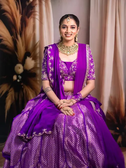 A classic seated bridal portrait. The composition is calm and elegant, allowing the intricate details of the purple lehenga and the gold jewelry to be the main focus.