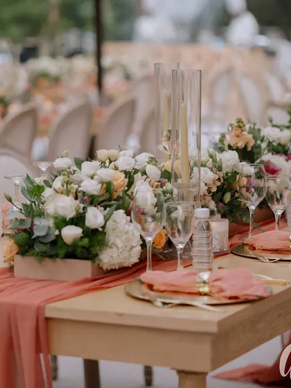 A close-up of the dining table setting, featuring peach table runners and napkins, gold-rimmed plates, and centerpieces of white and peach florals.