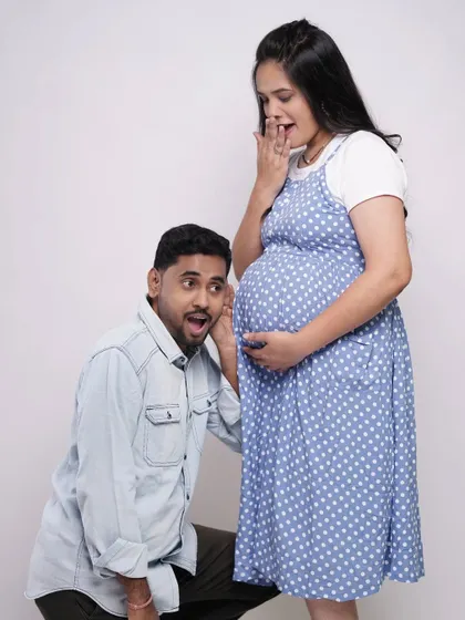 A fun and expressive studio shot. The father-to-be listens to the baby bump with a surprised look, while the mother-to-be reacts with a smile, capturing the playful side of their journey.