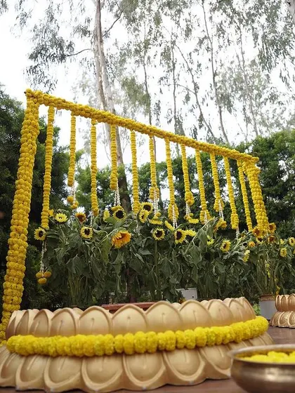 A beautiful Haldi setup designed to be one with nature. The backdrop is made of fresh sunflowers and marigold garlands, and the seating area is framed by large, traditional brass urlis and a lotus-shaped platform.