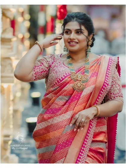 Varsha looking happy and radiant. Her makeup is soft and glowing, perfectly complementing her colorful pink and orange saree and traditional jewelry.