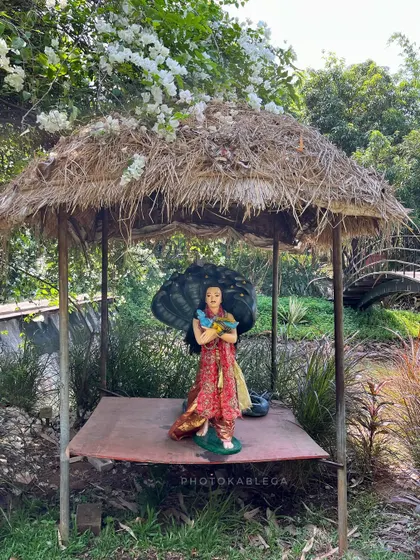 A statue of Krishna under a rustic thatched-roof shelter, surrounded by lush plants and white flowers.