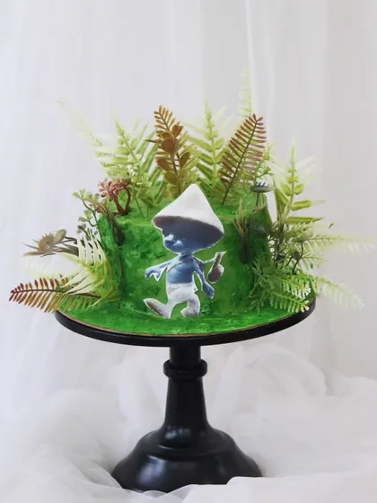 A unique and artistic Blue Smurf Cat cake set in a lush, green forest. The cake itself is textured to look like grass and is surrounded by realistic-looking ferns.