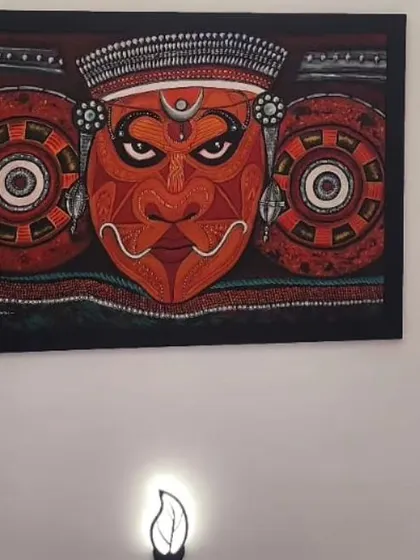 A powerful Theyyam mural hanging on a wall, illuminated by a small lamp below. This shows how the art can create a spiritual and dramatic focal point in a room.