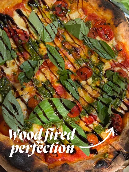 This is what we call wood-fired perfection. A beautiful pizza topped with fresh basil, cherry tomatoes, and a drizzle of balsamic glaze, showcasing the art of our pizza-making.