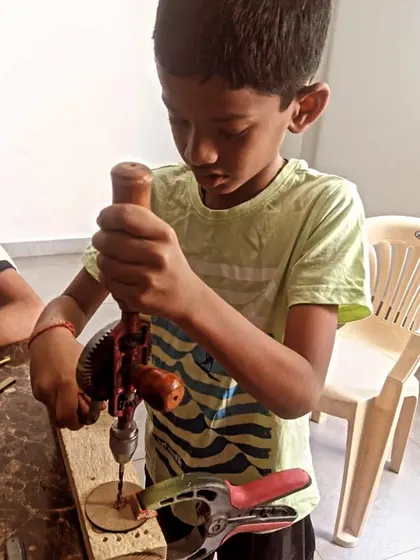 Focused on the task, this student uses a hand drill on a wooden disc, likely for a wheel on a car project.