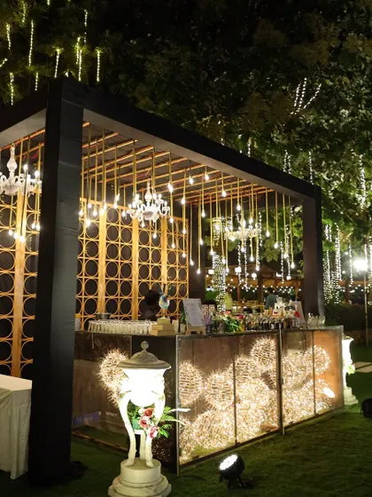 A stylish outdoor bar setup for a sangeet, featuring a modern black structure, intricate gold panels, and hanging Edison bulbs.