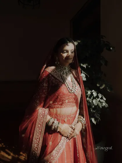 A dramatic bridal portrait with the bride standing in a sliver of sunlight, creating a beautiful play of light and shadow on her red lehenga.