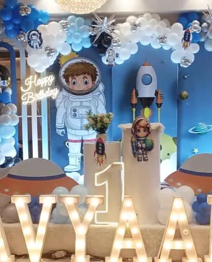 An out-of-this-world space-themed first birthday party. The decor includes an astronaut cutout, planet backdrops, and large "AVYAAN" marquee letters.