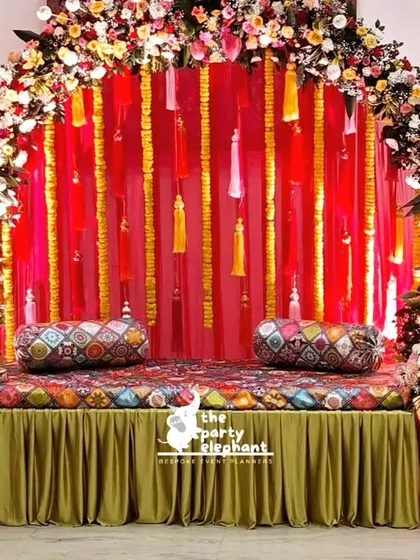 This vibrant backdrop was designed for a Mehendi ceremony. It features a rich red fabric with hanging tassels, framed by a lush arch of colorful flowers, and a decorated low seating area for the bride.