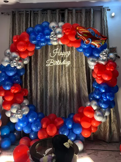 A circular balloon arch in red, blue, and silver for a car racing theme party at home. The arch is topped with a Lightning McQueen foil balloon and frames a "Happy Birthday" neon sign.