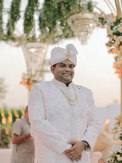 A portrait of the smiling groom, with the elegant details of the pastel floral mandap and hanging chandeliers softly blurred in the background.