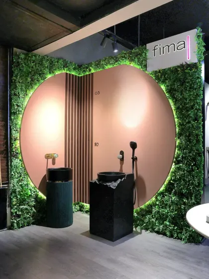 A creative display featuring pedestal sinks against a circular pink backdrop framed by a lush green wall, showcasing fixtures as art.