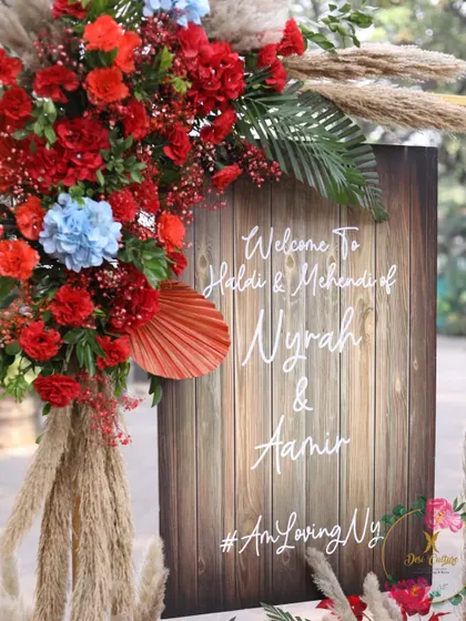 A rustic welcome sign for Nyrah and Aamir's Haldi and Mehendi, featuring a wooden texture and framed by pampas grass and red flowers.