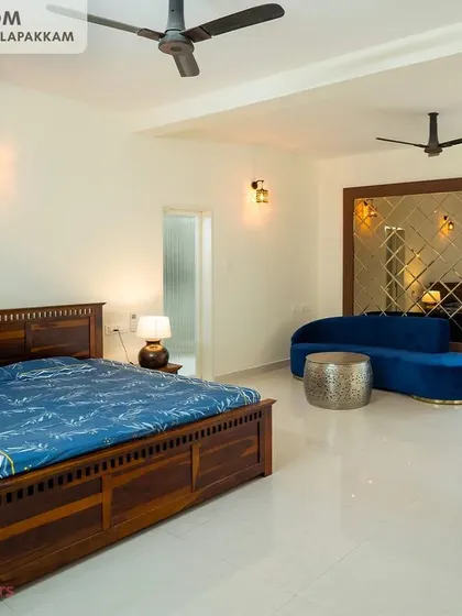 The master bedroom in the Sithalapakkam villa is a spacious retreat, featuring a traditional wooden bed, a modern blue sofa for lounging, and a large mirrored wall panel.