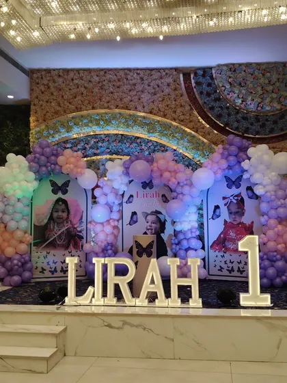 A grand stage for a first birthday party with a butterfly theme, featuring multiple photo backdrops and large light-up letters spelling the name "LIRAH".