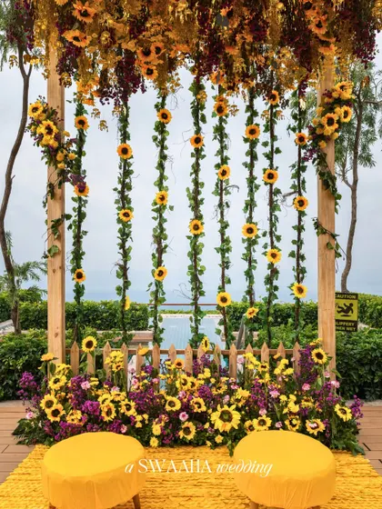 A creative photo backdrop made of a wooden frame with hanging sunflower garlands, set against a poolside view.