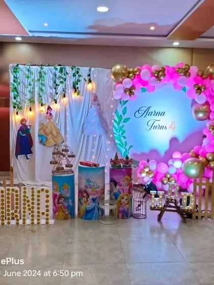 This Disney Princess party for Aarna's 4th birthday combines a curtain backdrop with a circular balloon arch. The setup includes princess cutouts, themed pedestals, and large marquee letters spelling "AARNA".
