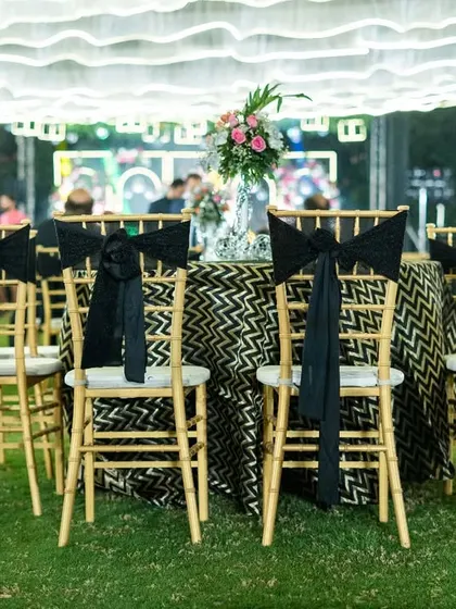 Guest seating at the Sangeet, featuring gold chiavari chairs with black sashes and tables with patterned black and gold linens, all under a ceiling of soft, draped fabric.