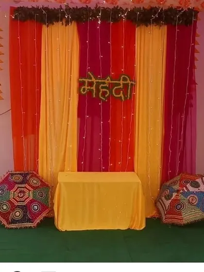 A simple Mehendi backdrop for a home function, using orange and yellow drapes, fairy lights, and decorative umbrellas.