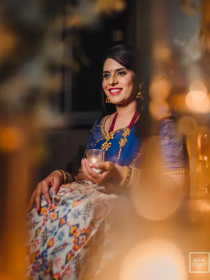 The bride smiles brightly, holding a candle, with a beautiful bokeh effect from other lights creating a magical atmosphere.