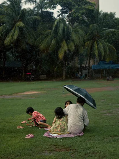 A close-up of the family having a picnic in the rain at Oval Maidan. This tighter shot focuses on the intimacy of the moment, with the father sheltering his children under the umbrella.