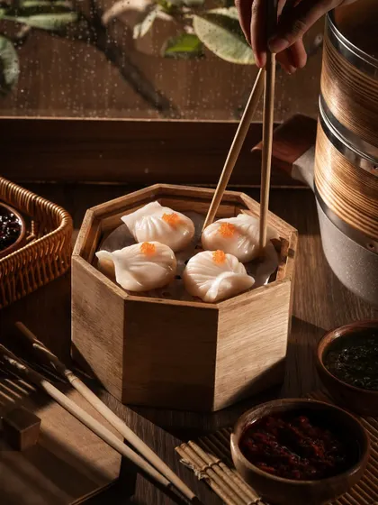 Using chopsticks to pick up a crystal shrimp dumpling (har gow). The lighting makes the translucent skin glow, showing the fresh filling inside.