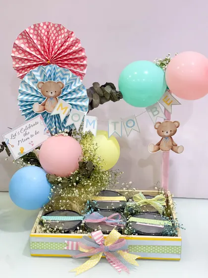 The full view of the mom-to-be food hamper, complete with a "Mom to Be" banner, teddy bear details, and colorful balloons, turning a meal delivery into a full-blown celebration.