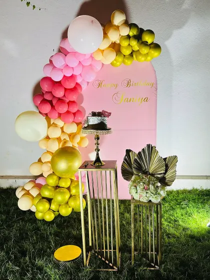 A simple yet beautiful outdoor setup on a lawn. This decoration includes a pink arched backdrop, a colorful balloon garland, and elegant golden stands for the cake and props.