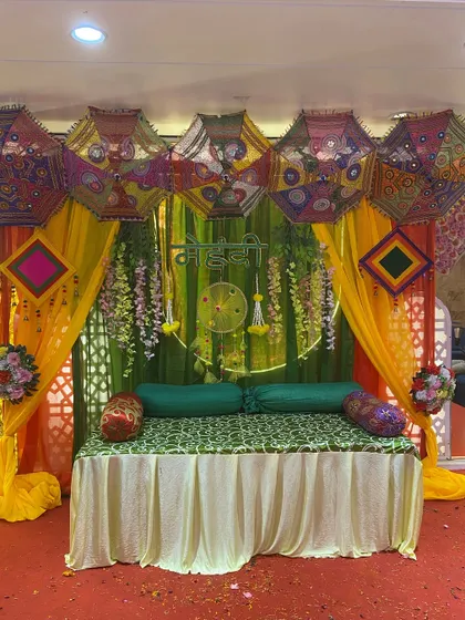 A lively mehndi setup with a green backdrop, colorful Rajasthani umbrellas, and hanging floral strings. The seating is kept simple to let the vibrant decor stand out.