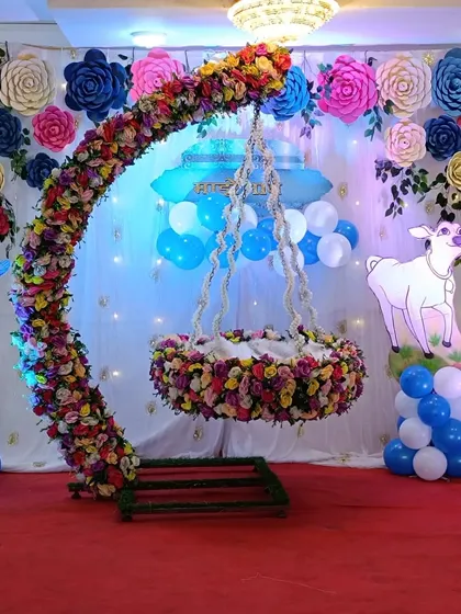 A Krishna-themed naming ceremony with a unique curved floral cradle. The backdrop features large paper flowers and cutouts of Krishna and a cow.