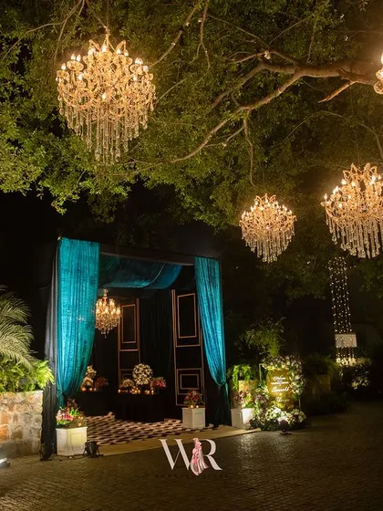 We love turning natural elements into features. Here, chandeliers hang like jewels from a majestic tree, creating a captivating entrance and setting a mood of whimsical elegance for this engagement night at Morbagh, Delhi.