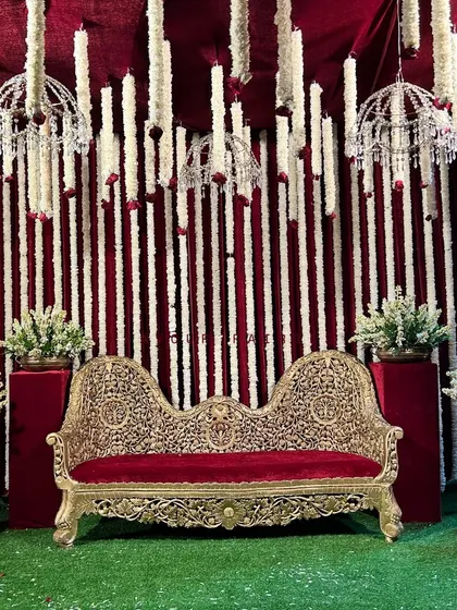 A full stage view of the traditional Godh Bharai ceremony. The ornate gold sofa sits on a green lawn against a backdrop of red velvet and hanging white floral garlands with crystal chandeliers.