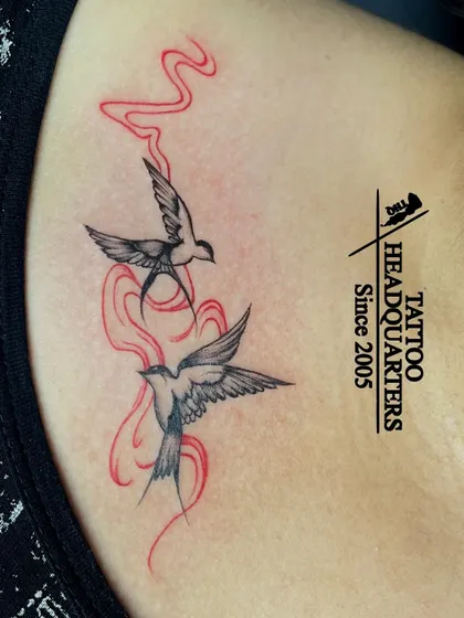 A tattoo of two swallows in flight, with red smoke-like trails, creating a dynamic and artistic piece.