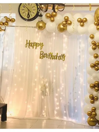 A simple yet elegant birthday decoration with a white and gold balloon garland, sheer curtain, and fairy lights.