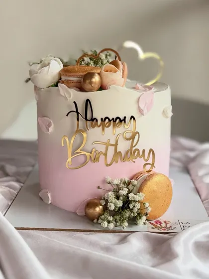 A pretty pink and white ombre cake decorated with macarons, baby's breath, and gold spheres. The gold heart topper adds a lovely finishing touch.