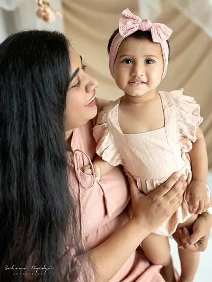 A mother and her baby girl, both looking at the camera with sweet smiles. The soft pink outfits and background create a lovely, gentle mood.