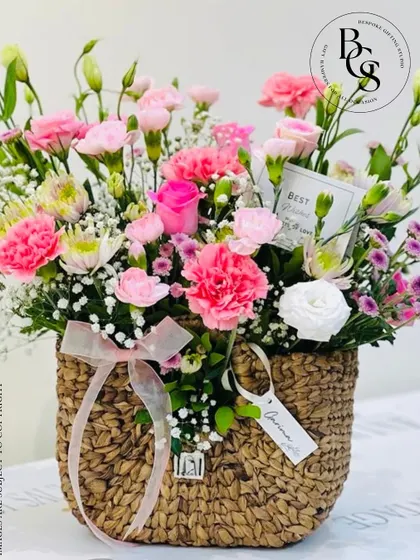 A charming purse-shaped wicker basket filled with a delicate arrangement of pink carnations and white accent flowers. This unique design is a whimsical and stylish way to gift flowers.