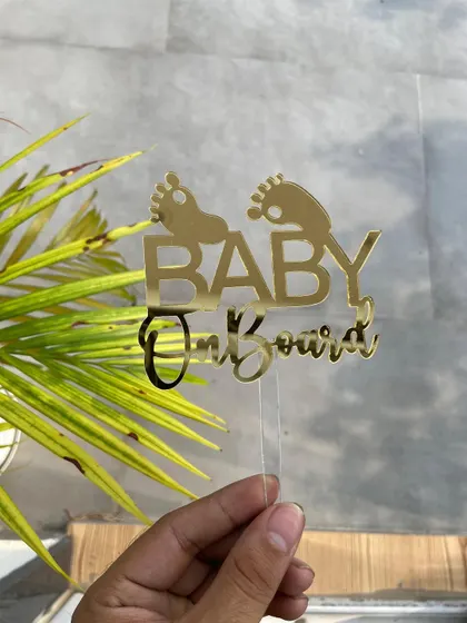 The "Baby on Board" topper held against a natural, green background, highlighting its vibrant gold color.