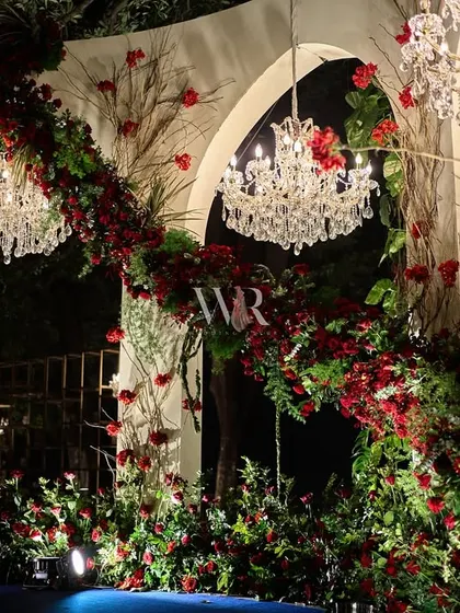 The engagement stage, where cascading red florals create a romantic arch over the couple's seating. The design feels organic and lush, as if the flowers are growing naturally over the structure.