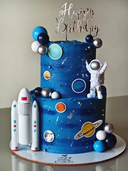 A two-tiered space-themed cake for a young astronaut's birthday. The galaxy effect was created with blue buttercream and silver dust, decorated with planets, rockets, and an astronaut topper.